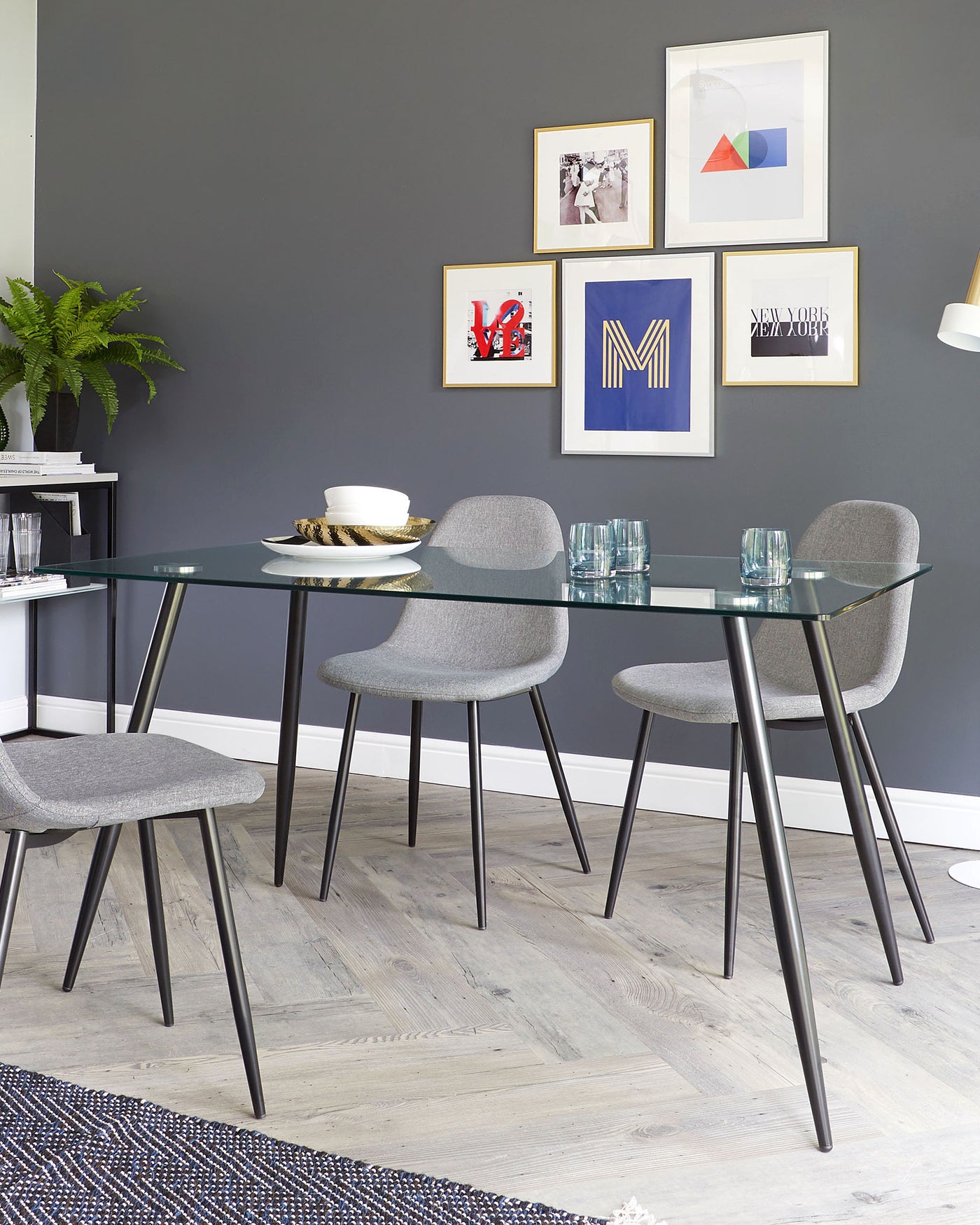 Modern dining area with a rectangular glass-top table on slim black metal legs and four matching grey upholstered chairs with sleek black frames. The setting reflects a contemporary minimalist style.