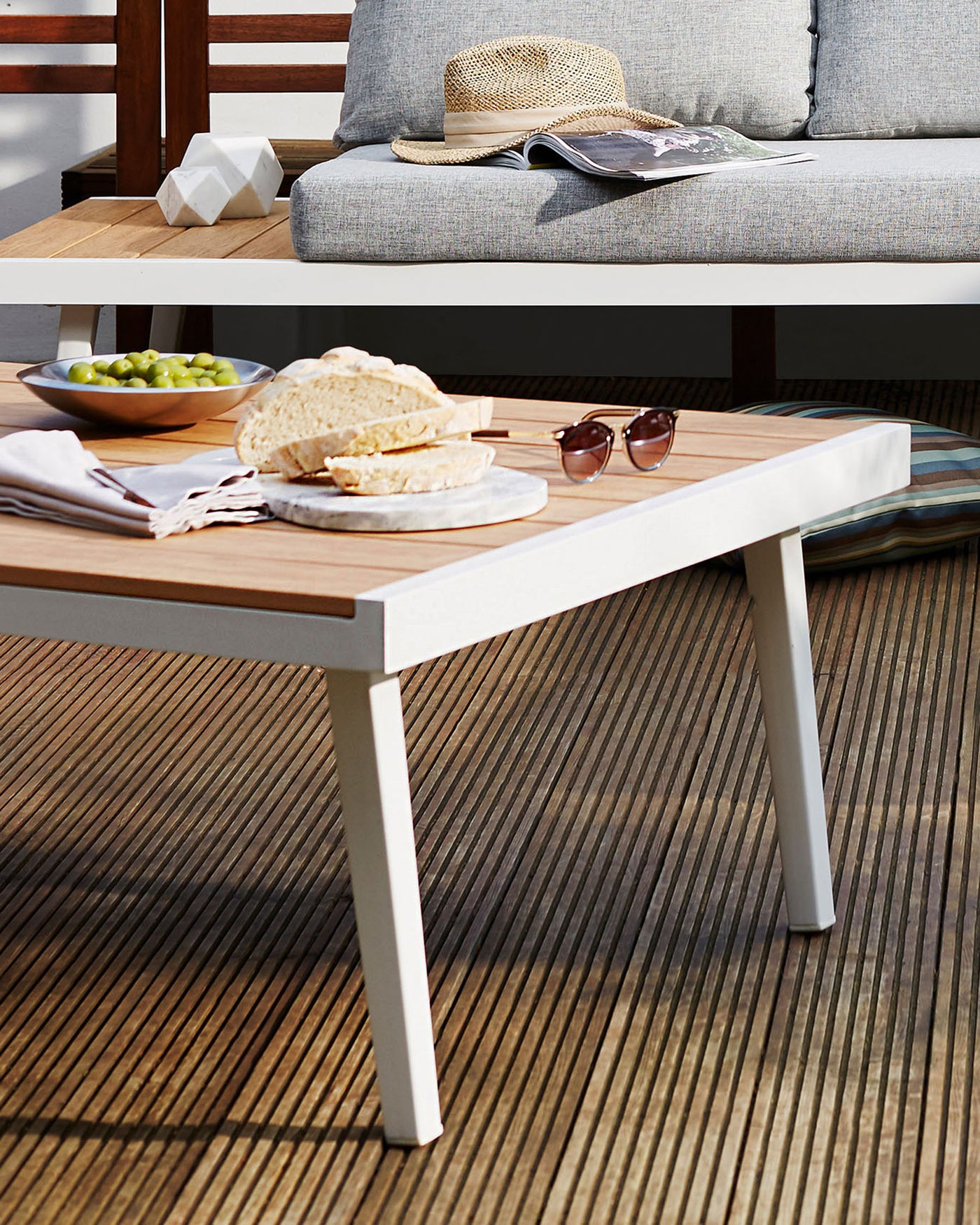 Modern outdoor two-tone wooden coffee table with white legs on a wooden deck, accompanied by a grey cushioned wooden sofa with decorative pillows, under natural sunlight.